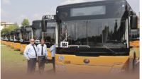 Flota nowoczesnych, ż&oacute;łto-czarnych autobus&oacute;w komunikacji miejskiej, zaparkowanych r&oacute;wno w długim rzędzie na placu. Na pierwszym planie, przed pojazdami, stoi grupa mężczyzn. Mają na sobie jasnoniebieskie koszule z kr&oacute;tkim rękawem, ciemne krawaty i ciemne spodnie, co sugeruje ubi&oacute;r służbowy kierowc&oacute;w transportu publicznego.