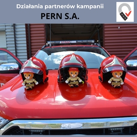 Baner promocyjny PERN S.A. w ramach kampanii &bdquo;Bezpiecznie do celu&rdquo;. Grafika zawiera nagł&oacute;wek informacyjny oraz zdjęcie z wnętrza garażu strażackiego, na kt&oacute;rym widać maskę czerwonego wozu z napisem &bdquo;STRAŻ&rdquo;. Ustawiono na niej trzy hełmy, a pod każdym z nich siedzi mała pluszowa maskotka rudego kota w miniaturowym stroju strażackim z logo &bdquo;PERN&rdquo;.